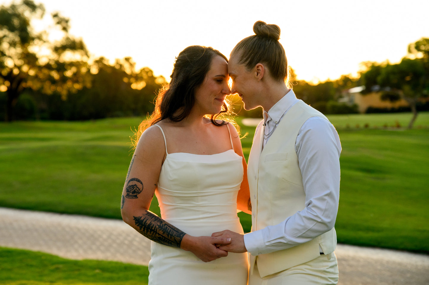 
                  
                    Two lesbian brides on their wedding day, embracing in a field. One bride wears the Club suit, a cream vest and pants with a white shirt, it is very dapper. The other bride wears a white dress.
                  
                