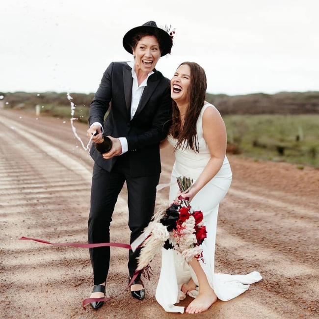 A queer wedding in the Australian outback. Two brides celebrate by popping a bottle of champagne. One bride is mas wearing a black tuxedo with peak lapels. One bride wears a white dress and is barefoot. 