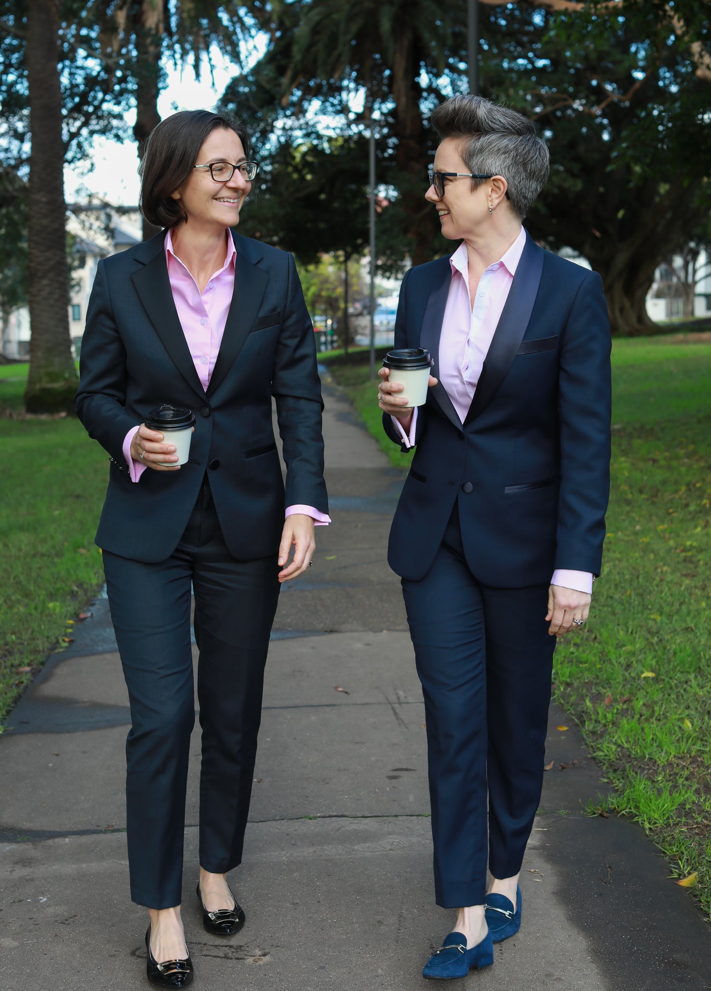 Two lesbian women walk through a park in pure wool tuxedo suits. 
