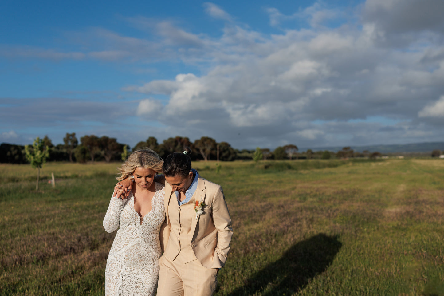 two lesbian women on their wedding day walking in a field, one bride wears a sand-coloured three-piece suit and a sky blue button-down shirt. The other bride wears a white dress.